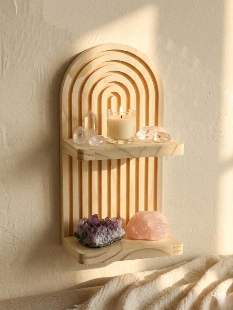 Puede incluir: Una estanter&iacute;a de pared de madera clara con una parte superior arqueada y dos estantes. El estante superior contiene una vela encendida en un vaso, cristales transparentes y un cristal de cuarzo. El estante inferior muestra una amatista p&uacute;rpura y un cristal de cuarzo rosa.