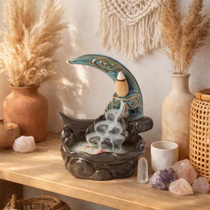 May include: A ceramic incense burner in the shape of a crescent moon, with a teal and gold finish. The burner sits on a wooden shelf, with smoke rising from it. Decorative items like crystals and vases surround the burner.