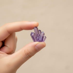 Könnte beinhalten: Ein Cluster aus kleinen, facettierten, lila Kristallen. Die Kristalle werden zwischen zwei Fingern vor einem neutralen Hintergrund gehalten. Die Kristalle haben ein durchscheinendes Aussehen und eine Vielzahl spitzer Formen.