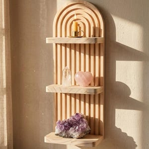 Puede incluir: Una estanter&iacute;a de pared de madera con tres niveles y una parte superior arqueada. Las estanter&iacute;as contienen una vela encendida, un cristal transparente, una piedra rosa en forma de coraz&oacute;n y un gran cristal de amatista p&uacute;rpura. La estanter&iacute;a es de madera clara.