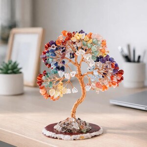 Op de afbeelding: Een decoratieve boomsculptuur met een koperkleurige draadstam en takken. De bladeren van de boom zijn gemaakt van kleurrijke edelstenen in tinten rood, oranje, geel, groen, blauw en paars. De boom staat op een ronde, donkerbruine basis.