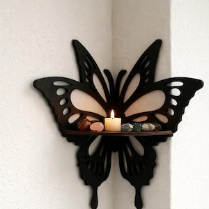 Puede incluir: Estantería de pared negra con forma de mariposa, con una vela encendida y pequeñas piedras. El diseño de la mariposa tiene detalles recortados y una balda de madera, sobre una pared blanca.
