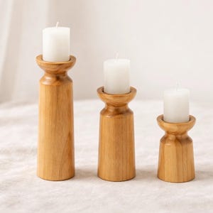 以下が含まれることがあります： 高さの異なる3つの木製キャンドルホルダー。それぞれに白いピラーキャンドルが立っています。キャンドルホルダーは明るい色の木材で作られ、旋盤加工が施されています。キャンドルは円筒形で、火は灯っていません。