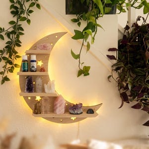 May include: A wooden crescent moon shelf with three tiers, illuminated by warm lights. The shelf is decorated with crystals and essential oil bottles. The design includes star cutouts and is mounted on a white wall, with hanging plants in the background.