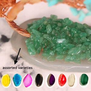 May include: A close-up of a decorative tree sculpture with a copper wire trunk and branches. The tree's leaves are made of small, green, polished stones. The base is a light gray oval with the text "assorted varieties" and a row of colorful stones.
