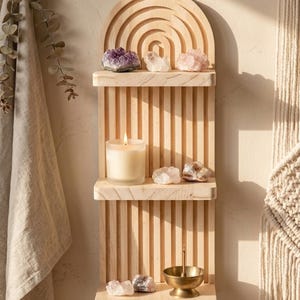 Puede incluir: Una estanter&iacute;a de madera de tres niveles con un dise&ntilde;o de arco iris en la parte superior. Las estanter&iacute;as contienen cristales, una vela encendida y un peque&ntilde;o cuenco de lat&oacute;n. La estanter&iacute;a est&aacute; montada en una pared de color claro, con un pa&ntilde;o colgante y decoraci&oacute;n de pared de macram&eacute;.