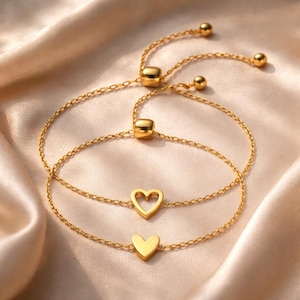 Puede incluir: Dos pulseras doradas con dijes de corazón. Una pulsera presenta un corazón macizo y la otra un recorte en forma de corazón. Las pulseras tienen cadenas ajustables y se exhiben sobre una superficie satinada.