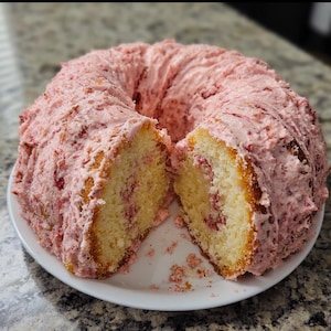 May include: A sliced, ring-shaped cake on a white plate. The cake has a light yellow interior with visible red fruit pieces. It is covered in a thick, pink frosting with visible fruit pieces. The cake is on a white plate.