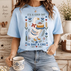 May include: A light blue t-shirt featuring a graphic of a white goose wearing a red bandana and holding a cake. The text reads IT'S A GOOD DAY TO BAKE A CAKE, surrounded by floral, wheat, and whisk illustrations in red, blue, and green colors.