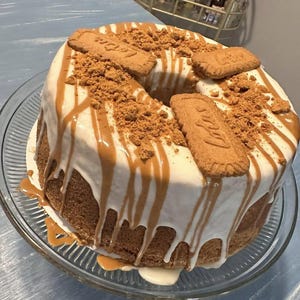 May include: A round bundt cake topped with white icing, drizzled with light brown caramel sauce, and garnished with crushed and whole Lotus Biscoff cookies. The cake sits on a clear glass pedestal stand, showcasing its textured golden-brown crumb.