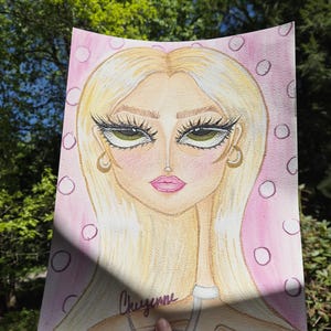 May include: Watercolor portrait of a woman with long blonde hair, green eyes, and pink lips. The artwork is on pink paper with white circles. The name "Cheyenne" is written in cursive. The artwork is held by a hand.