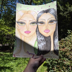 May include: A pencil drawing of two stylized female faces. One face has blonde hair and green eyes, the other has black hair and brown eyes. Both have pink lips and long eyelashes. The background is light green with pink polka dots. The artist's signature is visible.
