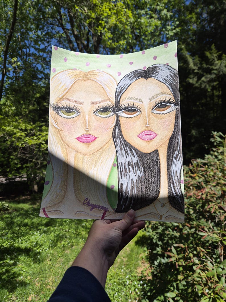 May include: A pencil drawing of two stylized female faces. One face has blonde hair and green eyes, the other has black hair and brown eyes. Both have pink lips and long eyelashes. The background is light green with pink polka dots. The artist's signature is visible.
