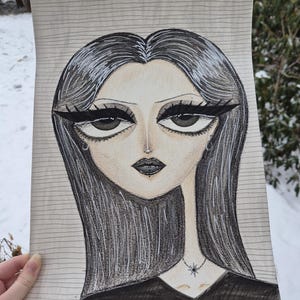 May include: A pencil drawing of a woman with long black hair and dramatic eye makeup. She wears a black top and a necklace with a star pendant. The artwork is on lined paper, held against a snowy outdoor background.