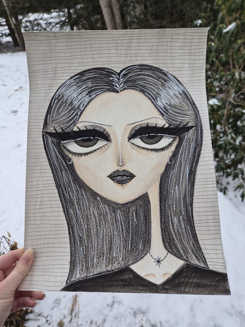 May include: A pencil drawing of a woman with long black hair and dramatic eye makeup. She wears a black top and a necklace with a star pendant. The artwork is on lined paper, held against a snowy outdoor background.