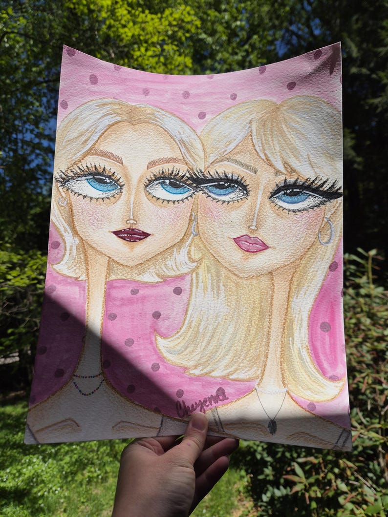 May include: Watercolor painting of two women with blonde hair and large blue eyes. The artwork features a pink background with polka dots. The women are wearing necklaces and the artist's signature is visible.