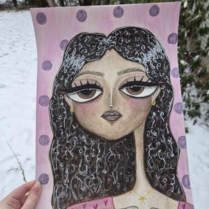 May include: An illustration of a woman with large brown eyes and dark curly hair. The artwork features a pink background with purple polka dots. The woman is wearing a pink top with a gold necklace. The artwork is held by a hand.