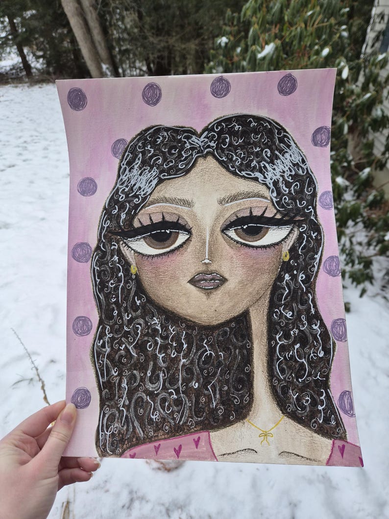 May include: An illustration of a woman with large brown eyes and dark curly hair. The artwork features a pink background with purple polka dots. The woman is wearing a pink top with a gold necklace. The artwork is held by a hand.
