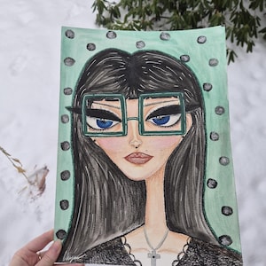 May include: A hand-drawn portrait of a woman with long black hair, blue eyes, and square green glasses. The artwork features a mint green background with black polka dots. The woman is wearing a black top and a silver cross necklace.