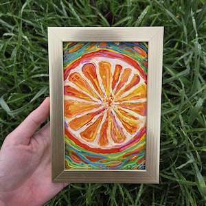 Könnte beinhalten: Gerahmtes Gemälde einer Orangenscheibe mit leuchtend orangefarbenen und gelben Segmenten. Das Kunstwerk hat einen farbenfrohen, abstrakten Hintergrund mit roten, blauen und grünen Farbtönen. Der goldene Rahmen verleiht diesem Kunstwerk zum Thema Obst einen Hauch von Eleganz.