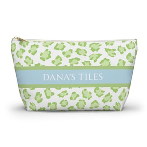 May include: A white cosmetic bag with a green leopard print pattern. A light blue band across the centre features the text "DANA'S TILES". The bag has a gold zip.