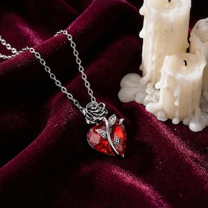 Puede incluir: Un collar plateado con un colgante rojo facetado en forma de corazón. Una rosa y hojas plateadas adornan el corazón. El collar se exhibe sobre una superficie de terciopelo rojo oscuro, con velas encendidas en el fondo.
