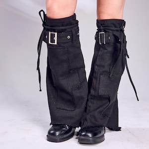 May include: Black leg warmers with a distressed, patched design. Each leg warmer features a silver buckle and tie closure. The bottom hem is frayed. The person is wearing black shoes.
