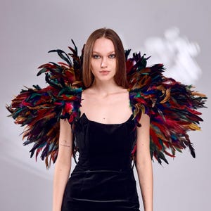 Könnte beinhalten: Eine Frau trägt ein schwarzes Samtkleid und einen auffälligen Schulteraufsatz aus Federn. Der Aufsatz besteht aus roten, grünen, blauen und braunen Federn, die ein farbenfrohes und auffälliges Accessoire bilden. Der Hintergrund ist ein neutrales Grau.