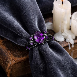 May include: Black metal ring with a large oval purple gemstone center and smaller purple accent stones. The ring has a floral design and is displayed on a dark gray velvet surface. The ring is a piece of jewelry.
