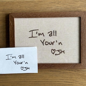 Op de afbeelding: Een ingelijst kunstwerk met de handgeschreven zin "I'm all Your'n" in zwarte inkt op een beige canvas. Een kleine witte notitie met dezelfde tekst en een hartsymbool is voor het frame geplaatst. Het frame is gemaakt van bruin hout.