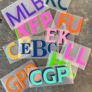 May include: A collection of heat transfer vinyl letters in various colors, including purple, pink, orange, blue, and green. The letters are arranged on clear sheets, with multiple letter combinations visible, such as MLB, KFP, and CGP.