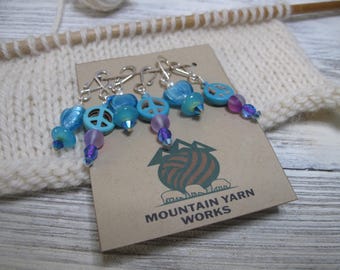 May include: A set of knitting stitch markers with blue peace sign charms and iridescent beads. The markers are attached to silver-colored clasps. A tag with the Mountain Yarn Works logo is visible, along with a knitted swatch and knitting needles.