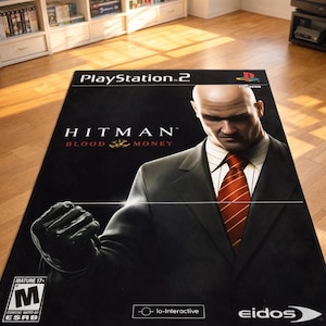 May include: A rectangular area rug featuring the PlayStation 2 video game cover art for Hitman Blood Money. The design displays a bald character in a black suit and red tie, the game title, the ESRB M rating, and the Io-Interactive and Eidos logos.