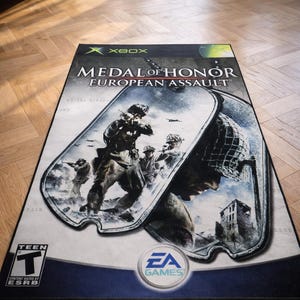 May include: A rectangular area rug featuring the Medal of Honor European Assault video game cover art for Xbox. The design displays soldiers in combat within a dog tag graphic, the EA Games logo, and an ESRB Teen rating symbol on a light background.