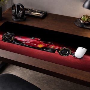 May include: A rectangular desk mat featuring a side profile of a red Ferrari Formula 1 race car against a dark background. The car displays sponsor logos including Ray-Ban, Ceva, and the Ferrari shield emblem. A white computer mouse sits on the mat.