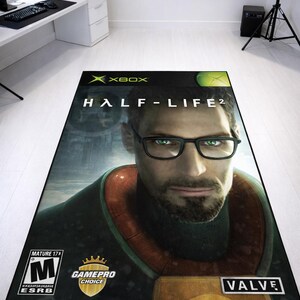 May include: A rectangular area rug featuring the Half-Life 2 video game cover art for Xbox. The design displays a portrait of a man with glasses, the Xbox logo, the Valve logo, a Mature 17+ ESRB rating, and a GamePro Choice gold shield emblem.
