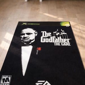 May include: A rectangular black rug featuring the cover art for The Godfather The Game for Xbox. The design includes a portrait of a man in a tuxedo with a red rose, the game title, the EA logo, an Xbox logo, and a Mature 17+ ESRB rating symbol.