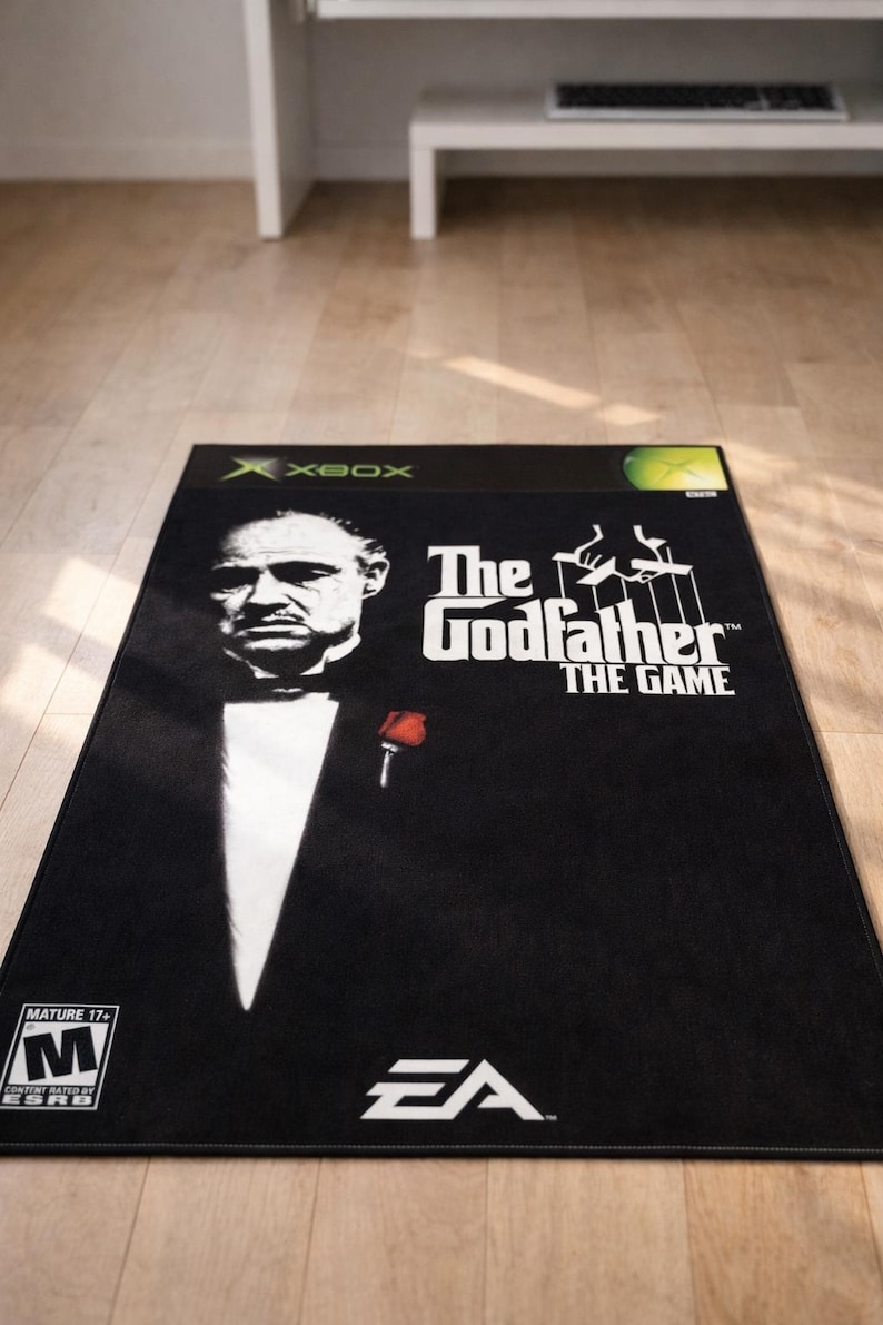 May include: A rectangular black rug featuring the cover art for The Godfather The Game for Xbox. The design includes a portrait of a man in a tuxedo with a red rose, the game title, the EA logo, an Xbox logo, and a Mature 17+ ESRB rating symbol.