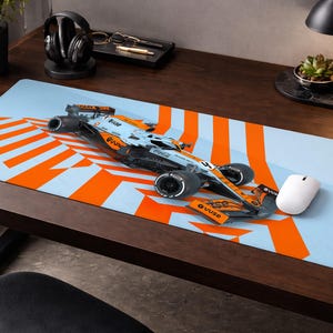 May include: A light blue and orange racing-themed desk mat with a Formula 1 race car model. The mat features a design of orange stripes and a white computer mouse. The desk mat is on a dark wooden desk.