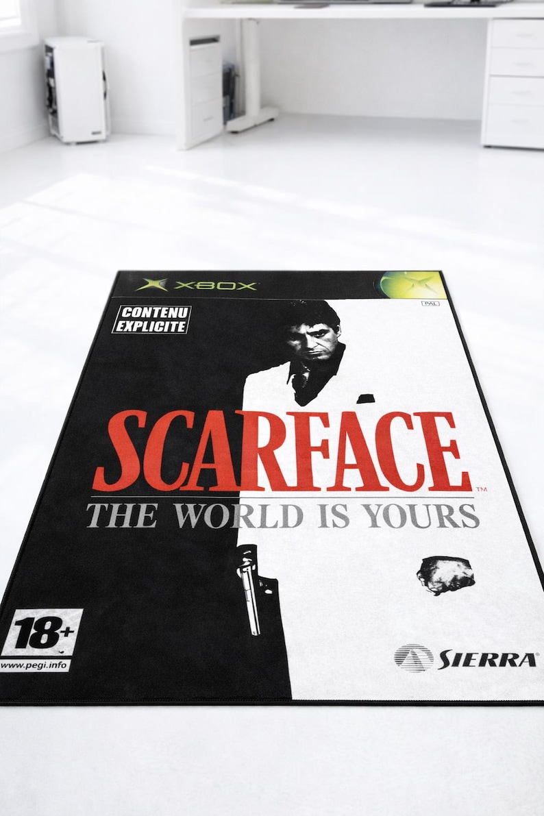 May include: A rectangular area rug featuring the Scarface: The World Is Yours video game cover art for Xbox. The design is split into black and white halves, displaying the title in red, the Sierra logo, an 18+ rating, and a Contenu Explicite label.