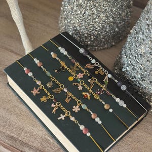 May include: A collection of handmade beaded bookmarks with gold-colored chains and charms. Each bookmark features a unique combination of beads, pearls, and decorative charms, including a starfish, turtle, and cross. The bookmarks are displayed on a closed book.