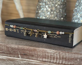 May include: A gold chain bookmark with various charms, including a cross, a clover, and letter charms, rests on a closed black book. The bookmark features beads in shades of pink, green, and white. The book's spine reads "Paolini."