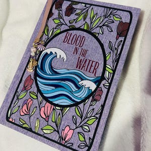 May include: A lavender journal with a floral design and the text "BLOOD IN THE WATER." The cover features a wave illustration in shades of blue. A pink ribbon bookmark with a charm of pearls, a shell, and a bell is attached.