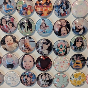 May include: A collection of round, clear glass magnets with various images. The magnets feature photos of people, pets, and decorative patterns. Some magnets have images of children, while others have floral and heart designs.