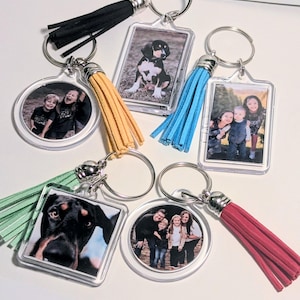 Könnte beinhalten: Eine Sammlung personalisierter Schlüsselanhänger mit Fotos, in verschiedenen Formen und mit farbenfrohen Quasten. Die Schlüsselanhänger zeigen Familienporträts und Tierfotos mit silbernen Schlüsselringen. Quasten sind in Schwarz, Gelb, Blau, Grün und Rot.