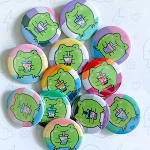 May include: A collection of colorful button pins featuring a green frog character holding a boba drink. The pins have various rainbow and pastel backgrounds. Text on the pins reads "Boba Frogs" and "Button Pins".