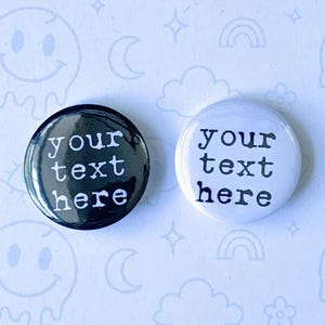May include: Two round button pins, one black and one white, each with the text "your text here" in a bold, sans-serif font. The words "BUTTON PINS" and "CUSTOM TEXT" are also visible. The background is light blue with repeating cartoon graphics.