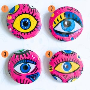 May include: Four round button pins with eye designs on a pink background. Each pin features a different eye design with blue, yellow, and white details. The text "BUTTON PINS" and "TOO AWARE FOR THIS" are also visible.