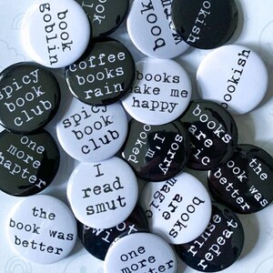 May include: A collection of black and white button pins with various book-related phrases like "spicy book club" and "books make me happy." The pins are scattered, with the text "COLLECT THEM ALL!" at the bottom and "MYSTERY BUTTON PINS" on the side.