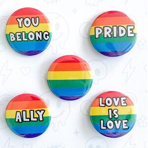 May include: A collection of rainbow-colored button pins with text including "YOU BELONG", "PRIDE", "ALLY", and "LOVE IS LOVE". The pins are labeled "BUTTON PINS" and "RAINBOW FLAGS".
